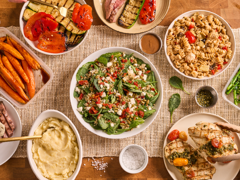 A spread of Feast & Fettle prepared meals, including salads, grilled vegetables, pasta, chicken, and sides arranged on a tabletop.